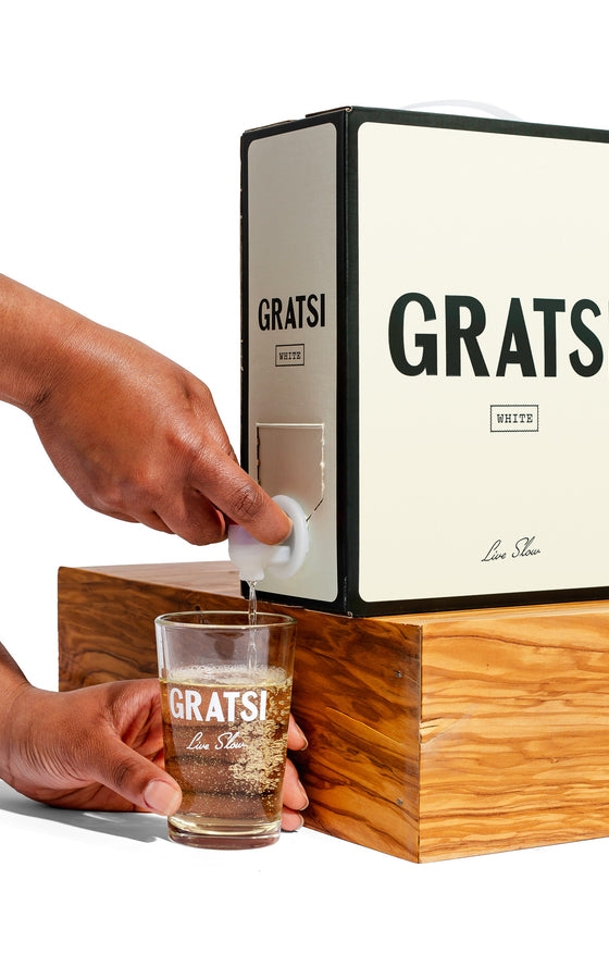 A person pours Gratsi White Wine, a zero sugar, low-sulfite boxed wine, from a large Gratsi box into a clear Gratsi-branded glass on a wooden block.