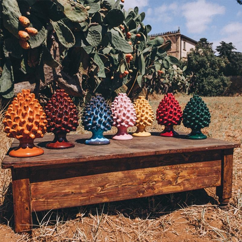 Seven colorful ceramic pinecone sculptures are displayed on a rustic wooden bench outdoors, with cactus plants and a building visible in the background.