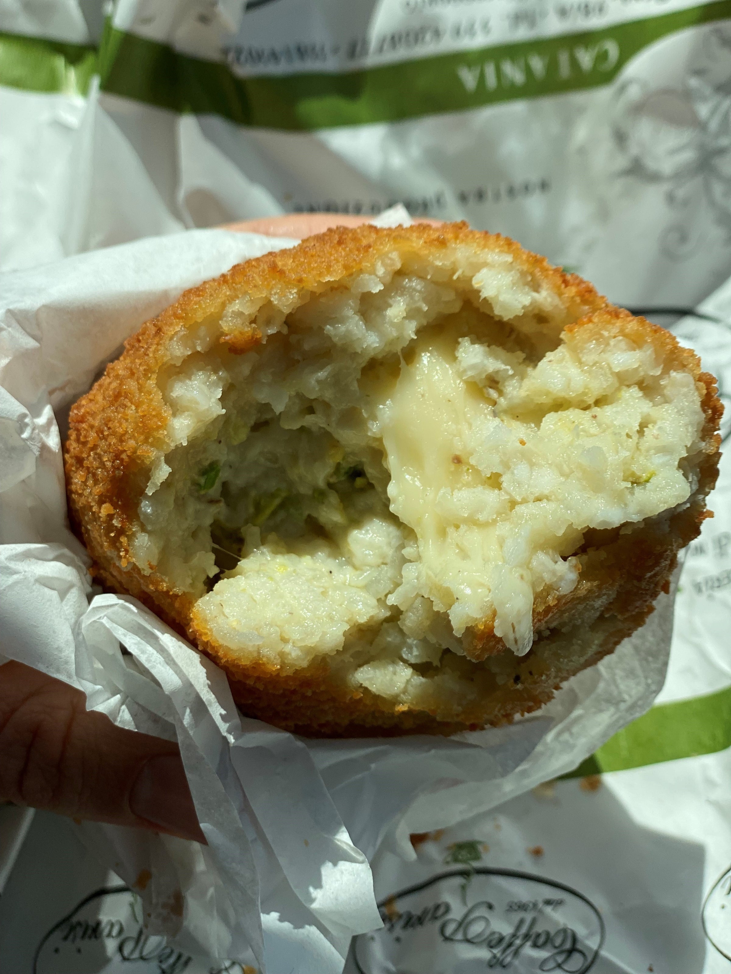 A close-up of a hand holding a half-eaten arancini, showing its crispy breaded rice exterior and creamy, cheesy filling inside. The arancini is wrapped in white paper.