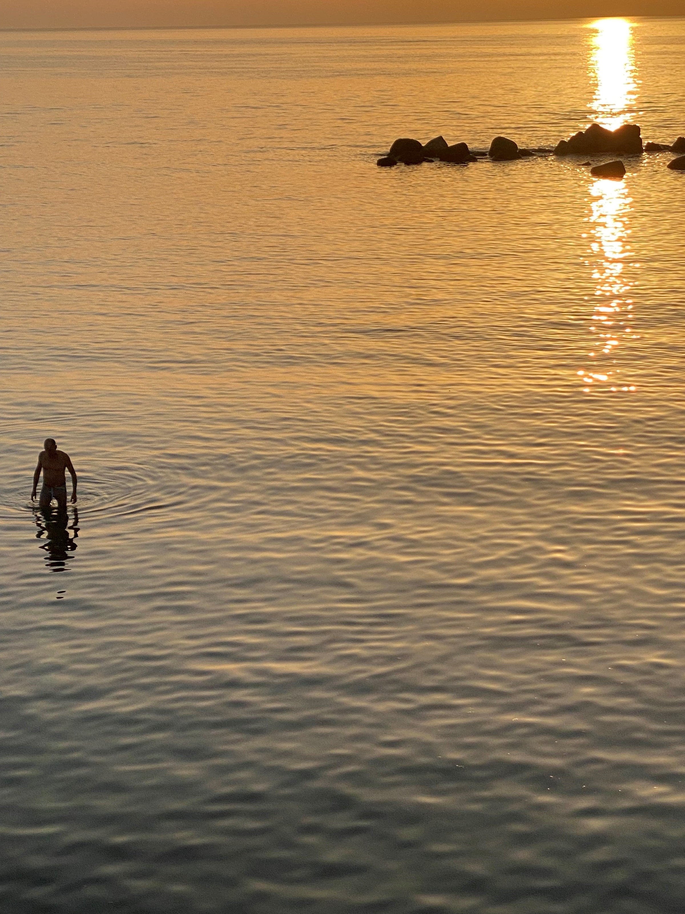 A person wades in calm water during sunset, with golden sunlight reflecting off the surface and rocks visible in the distance under an orange sky.