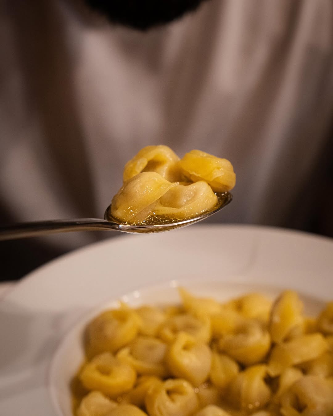 A close-up of a spoon holding several pieces of tortellini in broth, with a bowl of more tortellini in broth below. The background is blurred, focusing attention on the pasta.
