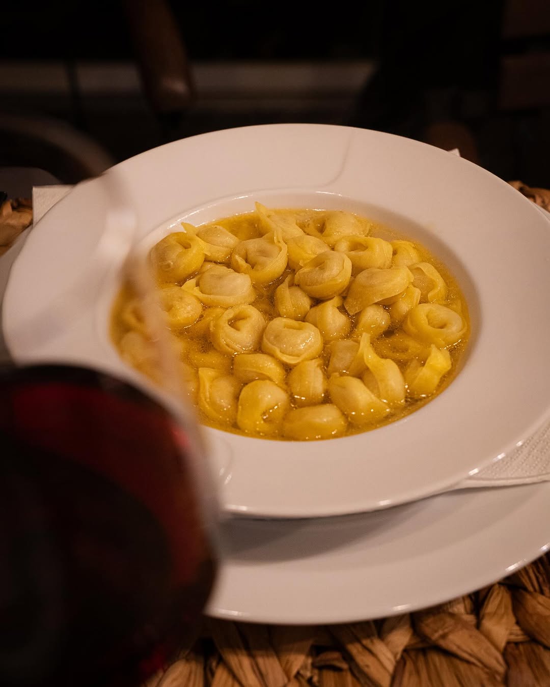 A white bowl filled with tortellini pasta in broth sits on a white plate, with a glass of red wine partially visible in the foreground.