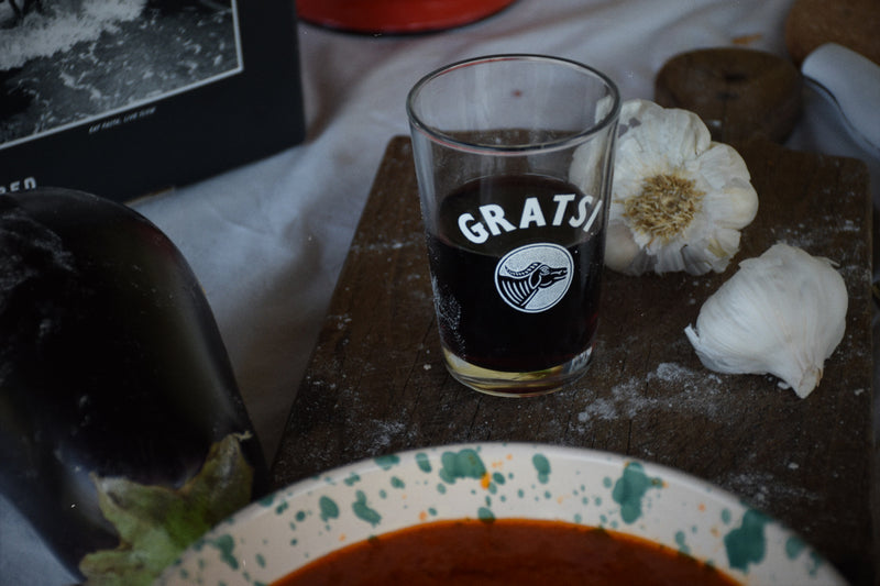 A glass with dark liquid labeled GRATS! sits on a wooden board next to garlic cloves and a bulb, with an eggplant and a bowl of red sauce in the foreground.