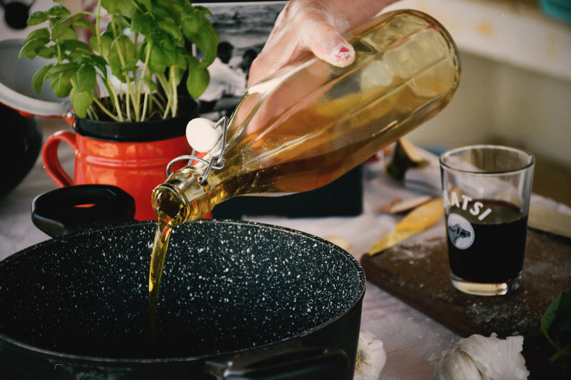 A hand pours oil from a glass bottle into a black speckled pot. A red mug with a basil plant, a glass of dark liquid, and cloves of garlic are on the table nearby.