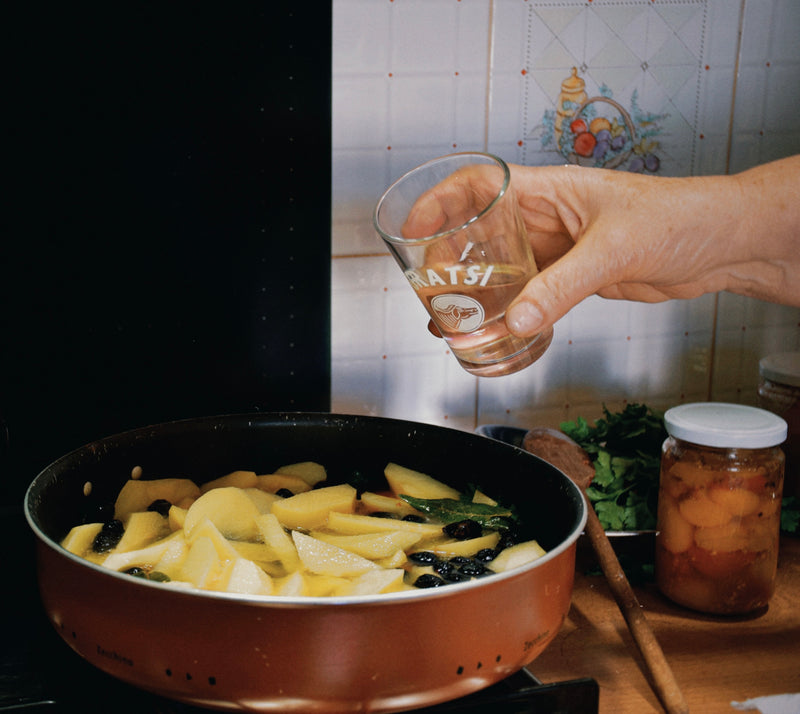 A hand pours liquid from a small glass into a pan filled with sliced apples and berries on a stove. A jar of fruit preserves, chopped herbs, and a wooden spoon sit nearby on the kitchen counter.