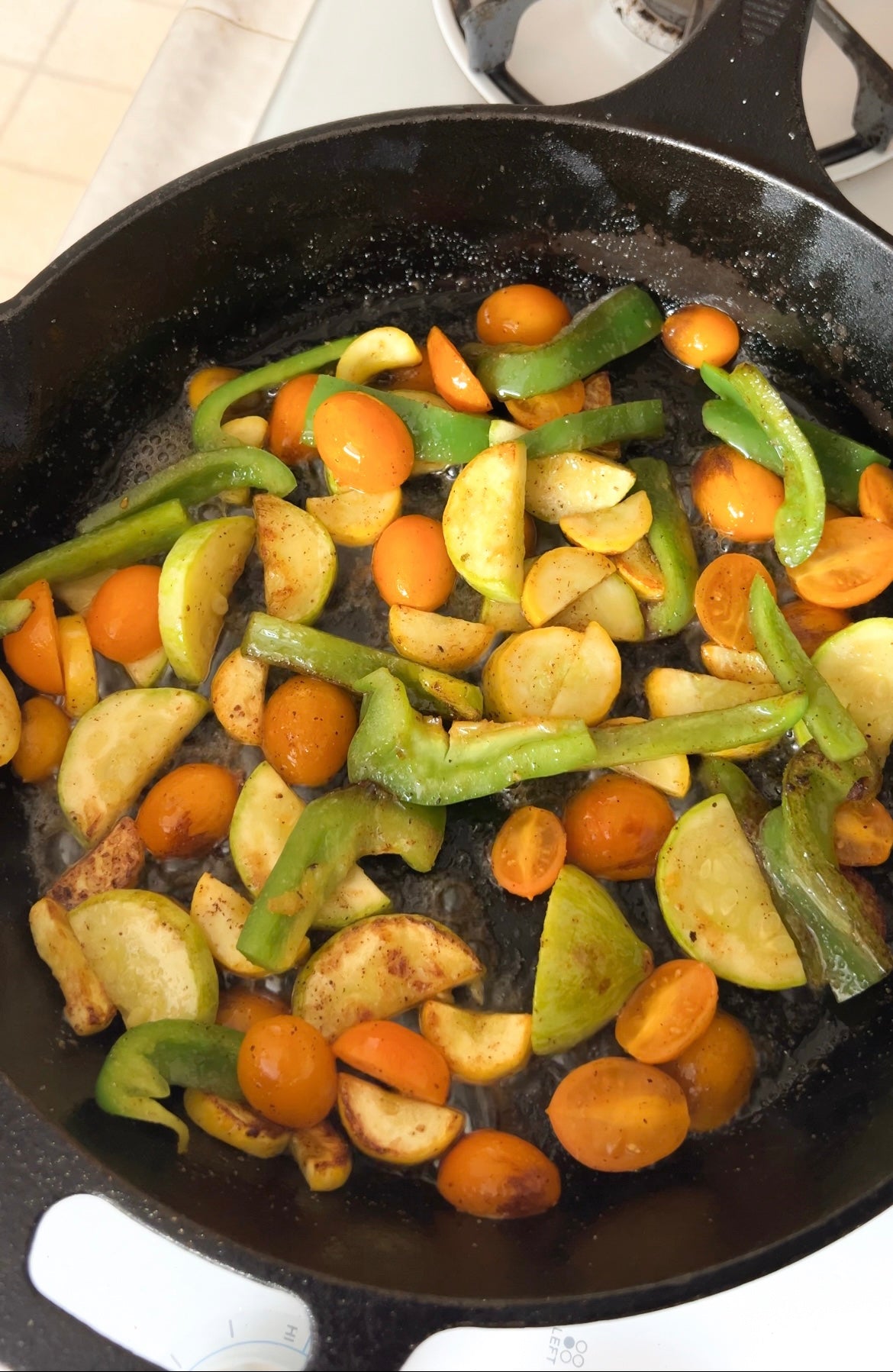 A cast-iron skillet on a stove contains sautéed vegetables, including sliced green bell peppers, yellow squash, and halved orange cherry tomatoes. The vegetables appear lightly cooked and seasoned.