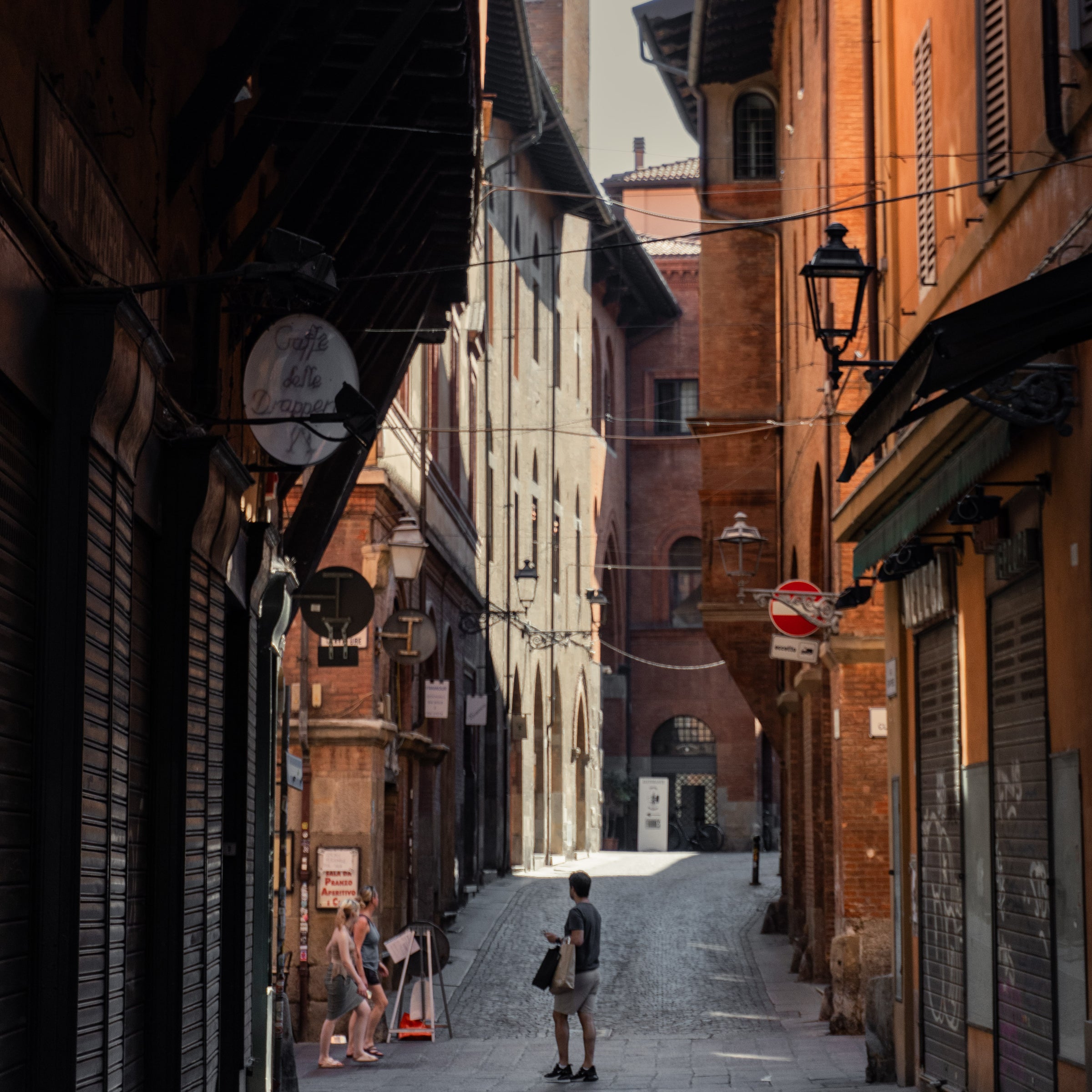 A narrow, cobblestone street lined with old brick buildings, shuttered shopfronts, and vintage street lamps. Three people walk and talk near a cafe sign as sunlight filters through the alley.