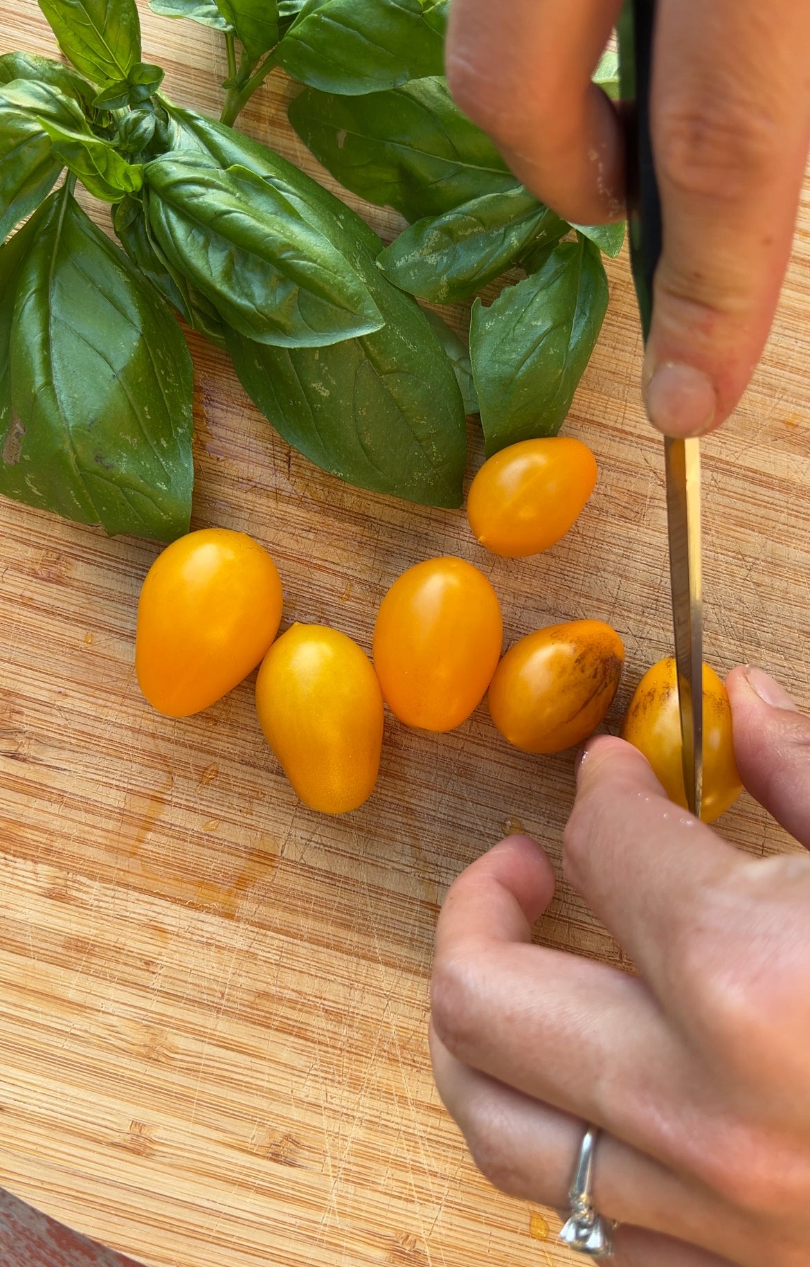 A hand with a ring slices a yellow grape tomato on a wooden cutting board, next to a bunch of fresh basil leaves and more whole yellow tomatoes.