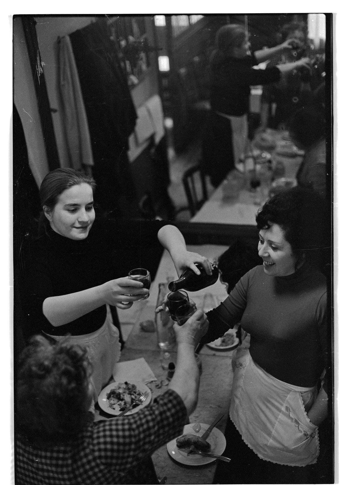Three women, some wearing aprons, smile and raise glasses in a toast at a crowded dining table filled with plates of food in a busy, lively restaurant.