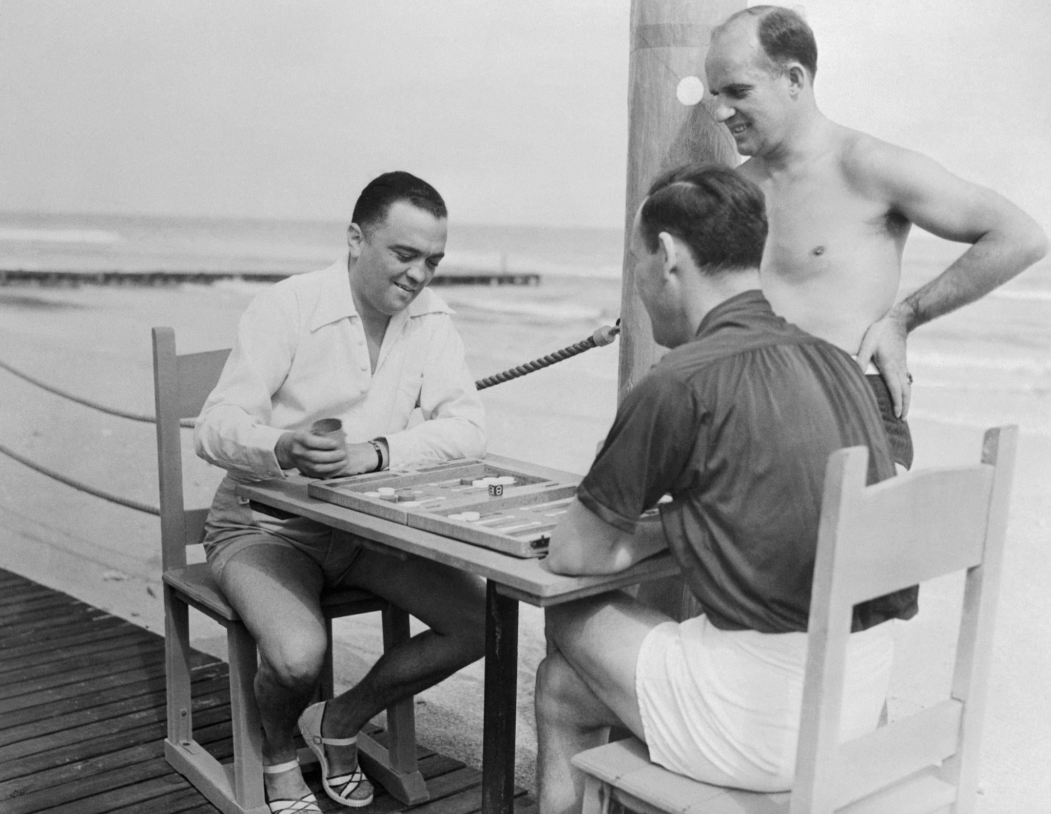 Three men in casual beachwear play a board game at a small table by the seaside; two are seated, one stands shirtless, all appear relaxed and engaged near the beach with waves in the background.