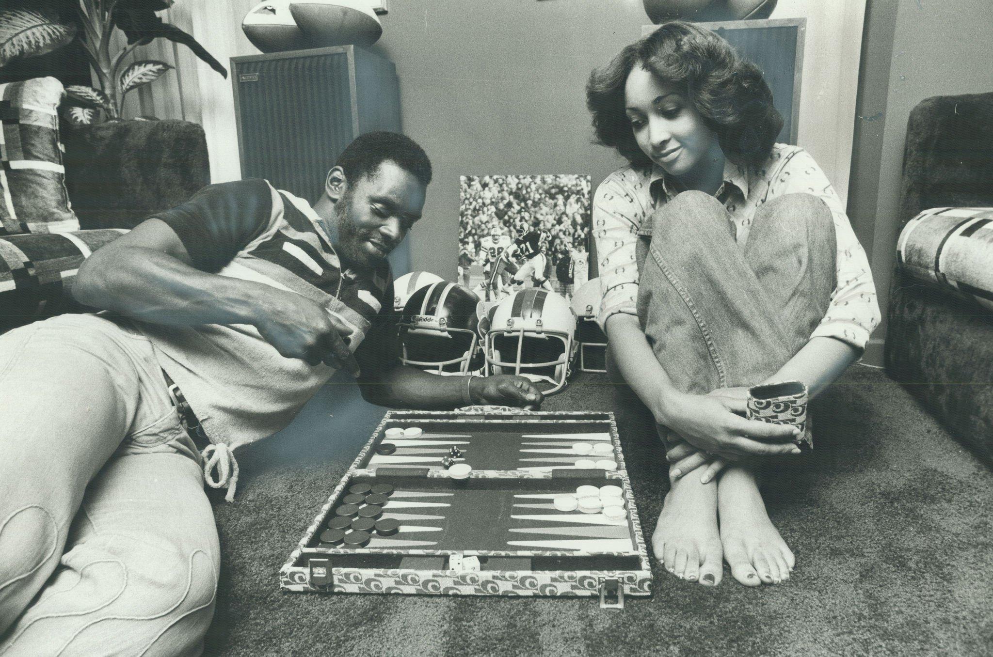 A man and a woman sit on the carpeted floor playing backgammon. Two football helmets are behind the board, and both appear relaxed and smiling. The room is decorated with plants, speakers, and sports memorabilia.