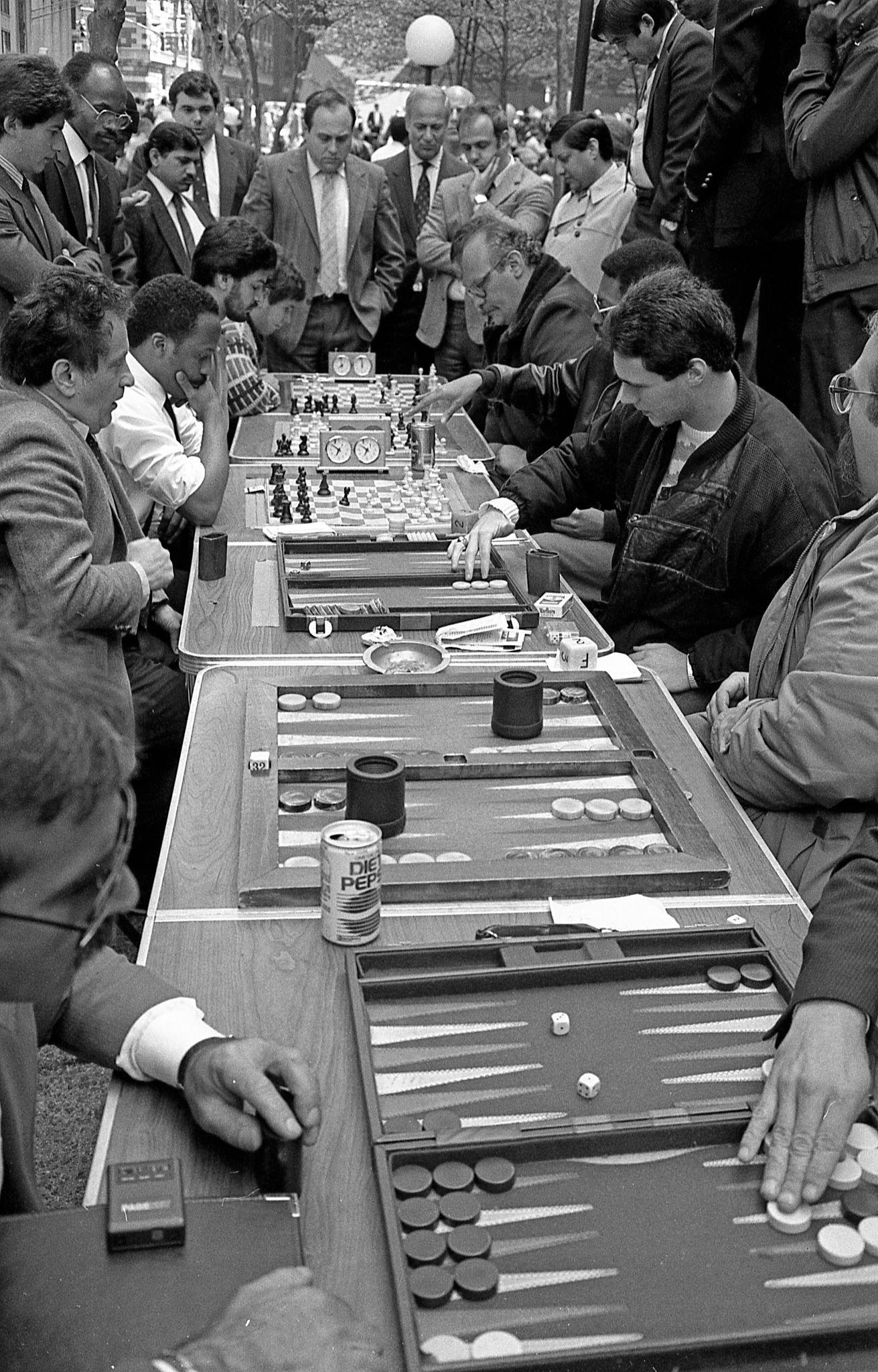 A group of men are gathered outdoors around tables playing chess and backgammon, with a crowd watching. The atmosphere is focused and intense. There is a can of Diet Pepsi on one table. The scene appears lively and crowded.
