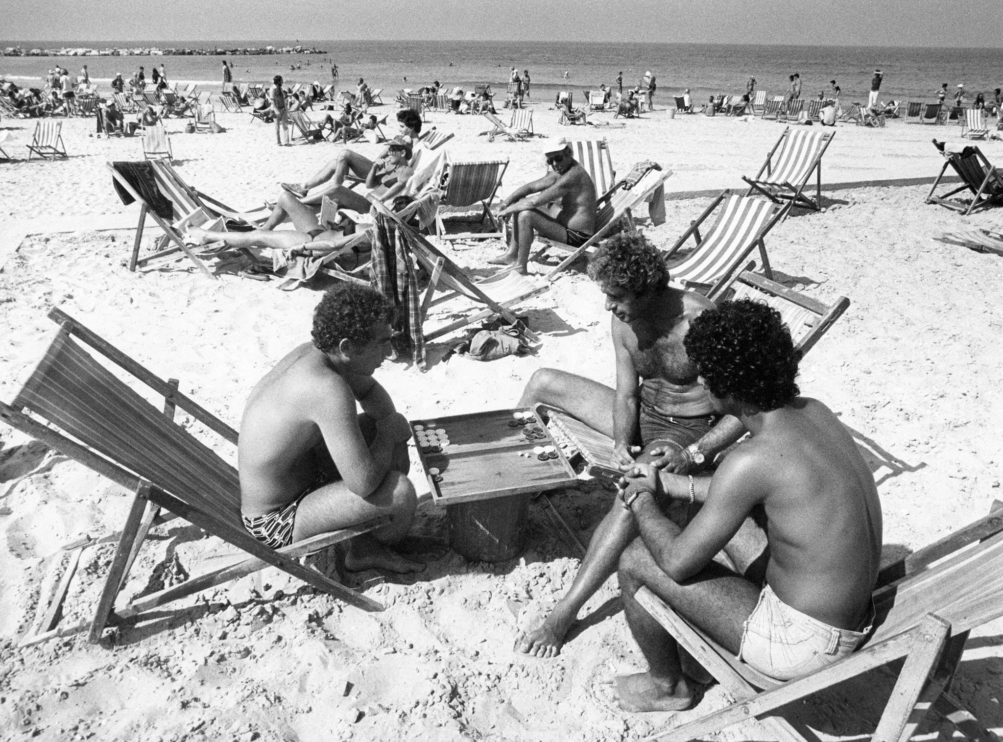 Three men sit on deck chairs playing a board game on a sandy beach. Other people relax in deck chairs and sunbathe in the background near the sea under a clear sky. The image is in black and white.