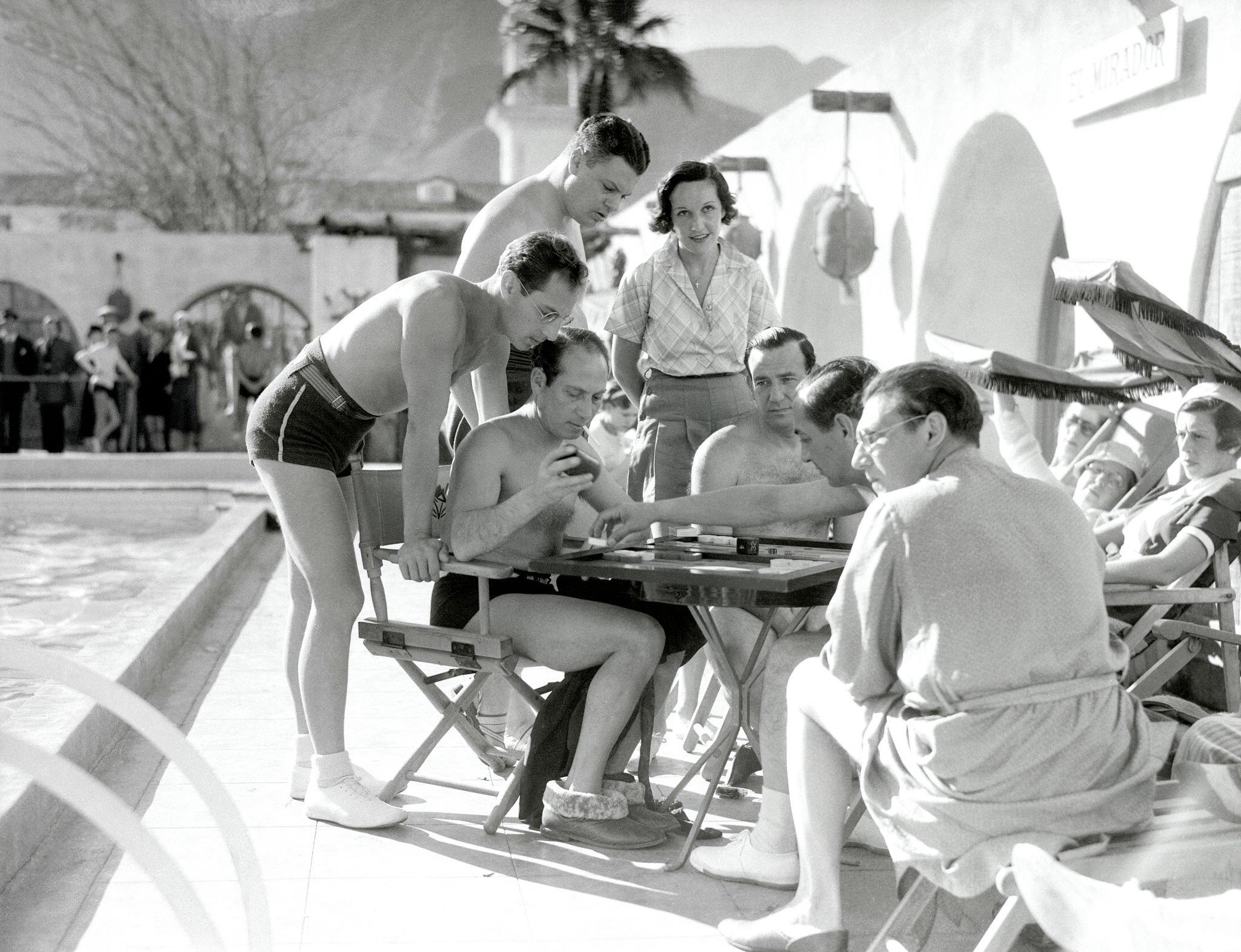 A group of men in swimwear gather around a table playing a board game by a pool, while a woman and others look on in a sunny outdoor setting. Palm trees and mountains are visible in the background.