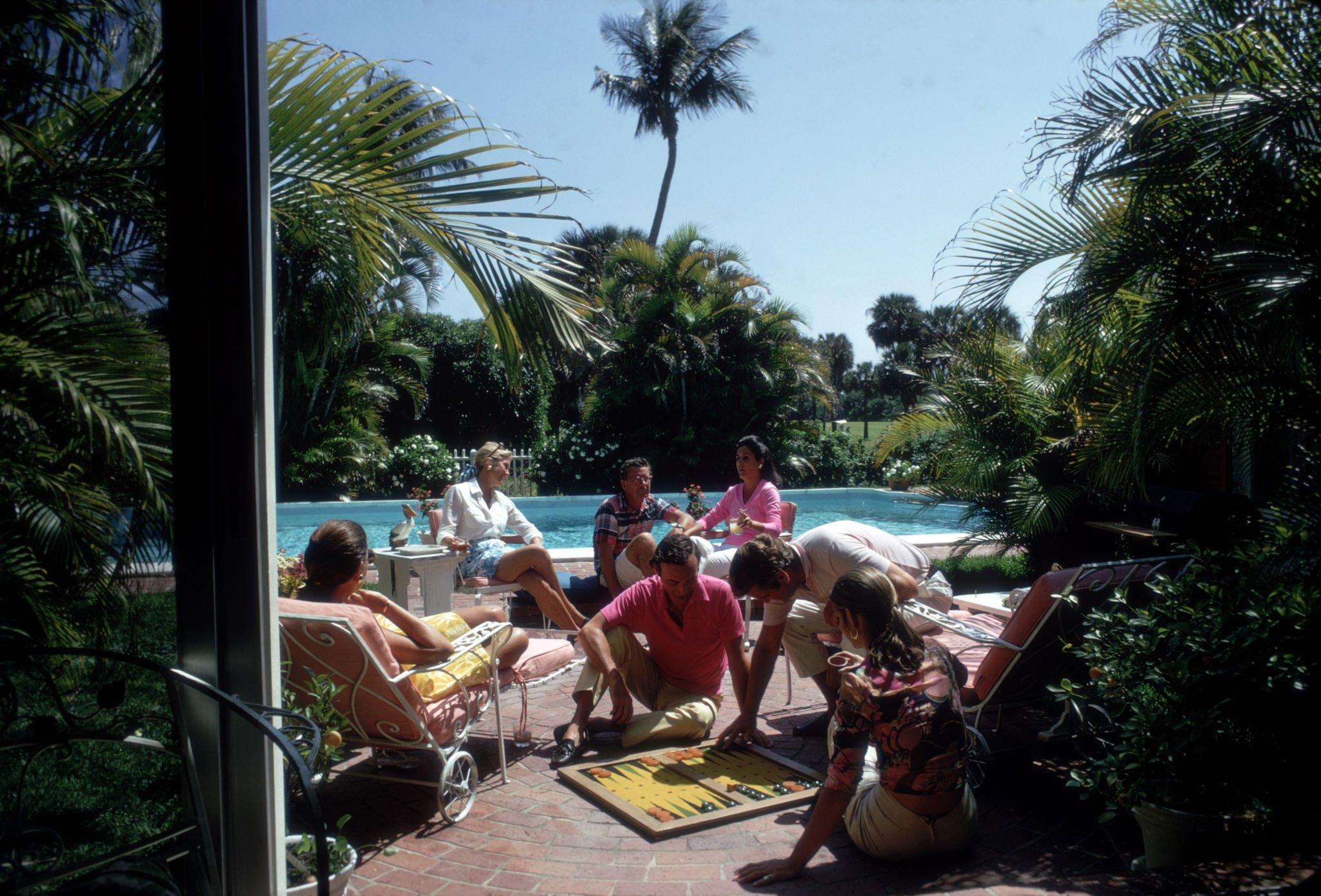 A group of people relax beside a swimming pool, some lounging on chairs, others playing a board game on the ground, with lush greenery and palm trees in the background under a bright sky.