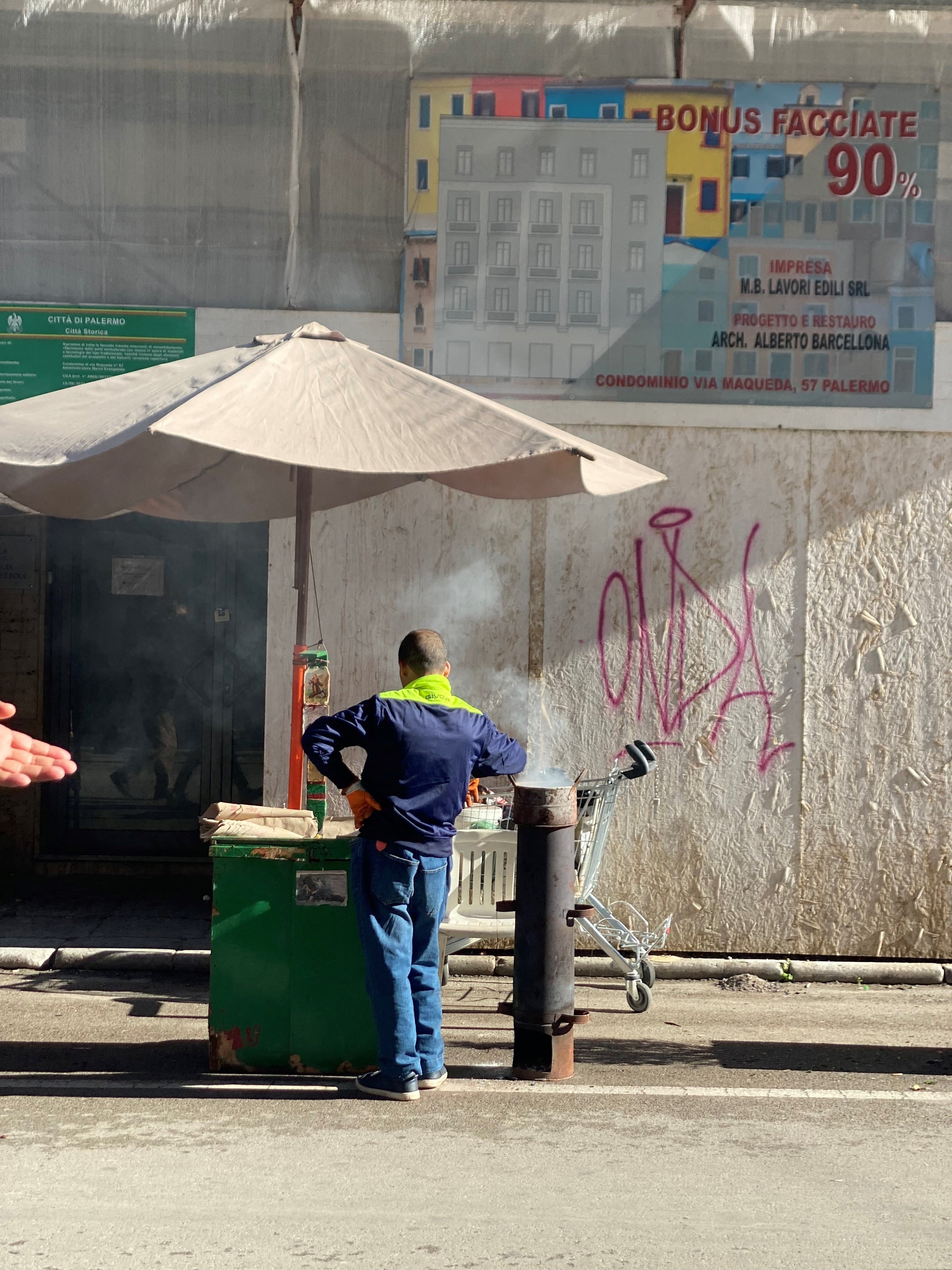A person stands by a green street food cart with smoke rising, under a large beige umbrella on a sunny day. A shopping cart and a graffitied wall are in the background.