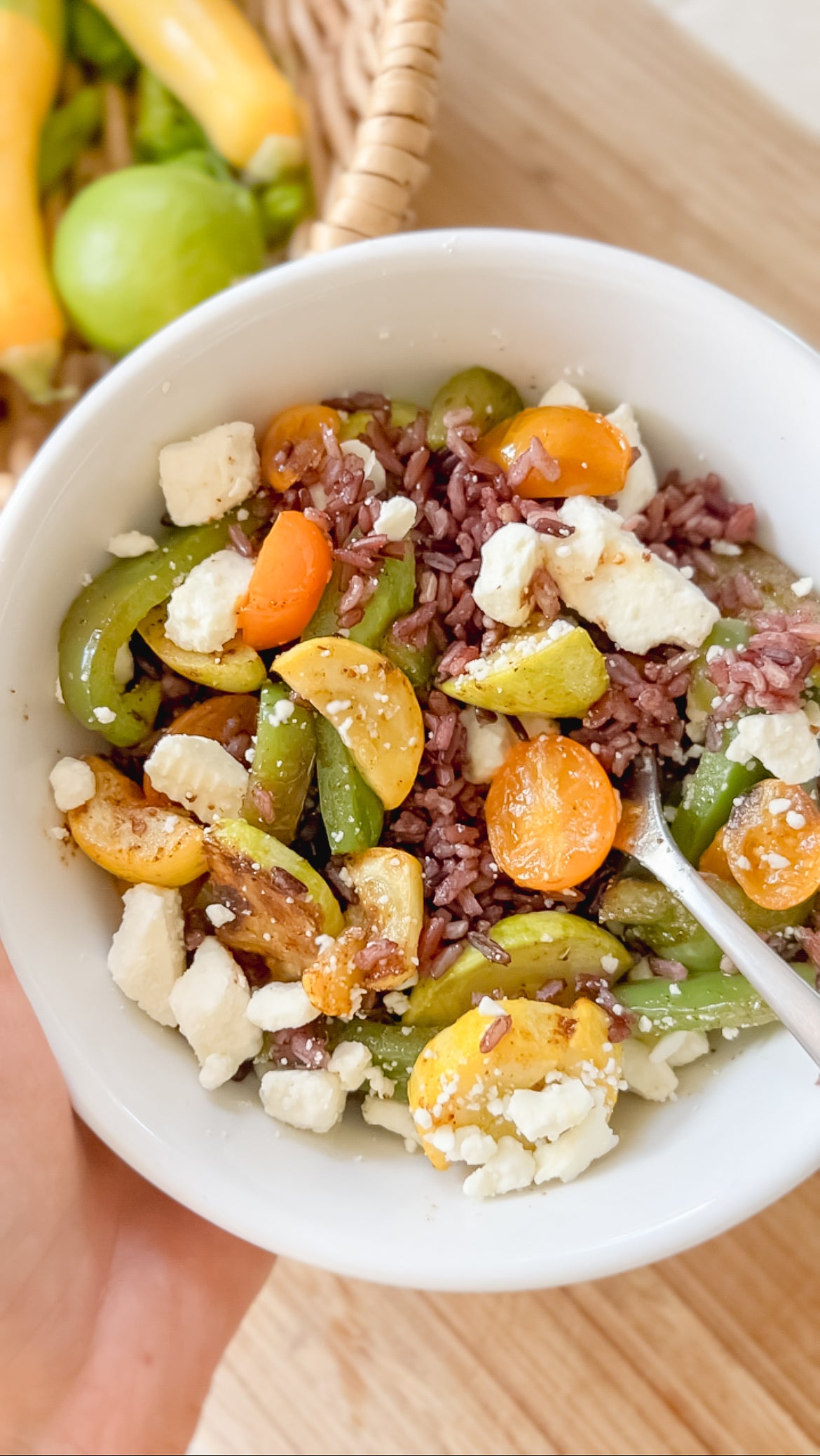 A white bowl filled with purple rice, sliced cherry tomatoes, green bell peppers, yellow squash, and crumbled feta cheese, with a fork inside. A basket of fresh vegetables is visible in the background.