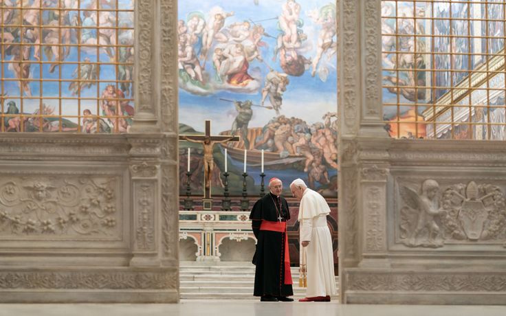 Two Catholic clergy, one in white and one in black and red, stand and talk inside a grand chapel with ornate marble columns and a large mural-covered ceiling, including Michelangelo’s “The Last Judgment,” visible in the background.