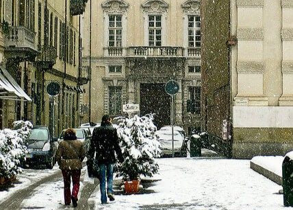 Two people walk down a snowy city street lined with parked cars and buildings. Snow is falling, covering the ground, trees, and rooftops. The scene appears calm and wintry.