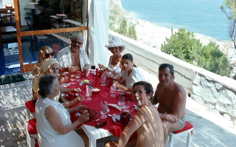 A group of eight people in swimsuits sit around a red tablecloth sharing a meal outdoors on a sunny patio overlooking the sea, with trees and rocky coastline in the background.
