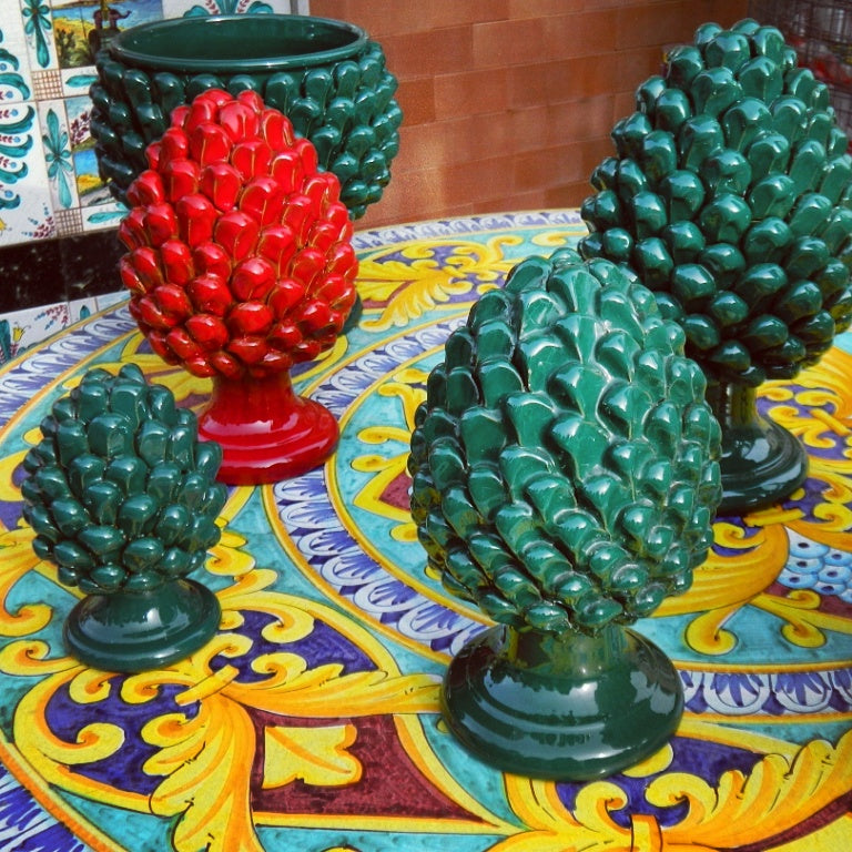 Colorful ceramic pine cone sculptures in green and red are displayed on a round, ornate table with a vibrant, decorative yellow, blue, and orange pattern. A tiled wall is visible in the background.