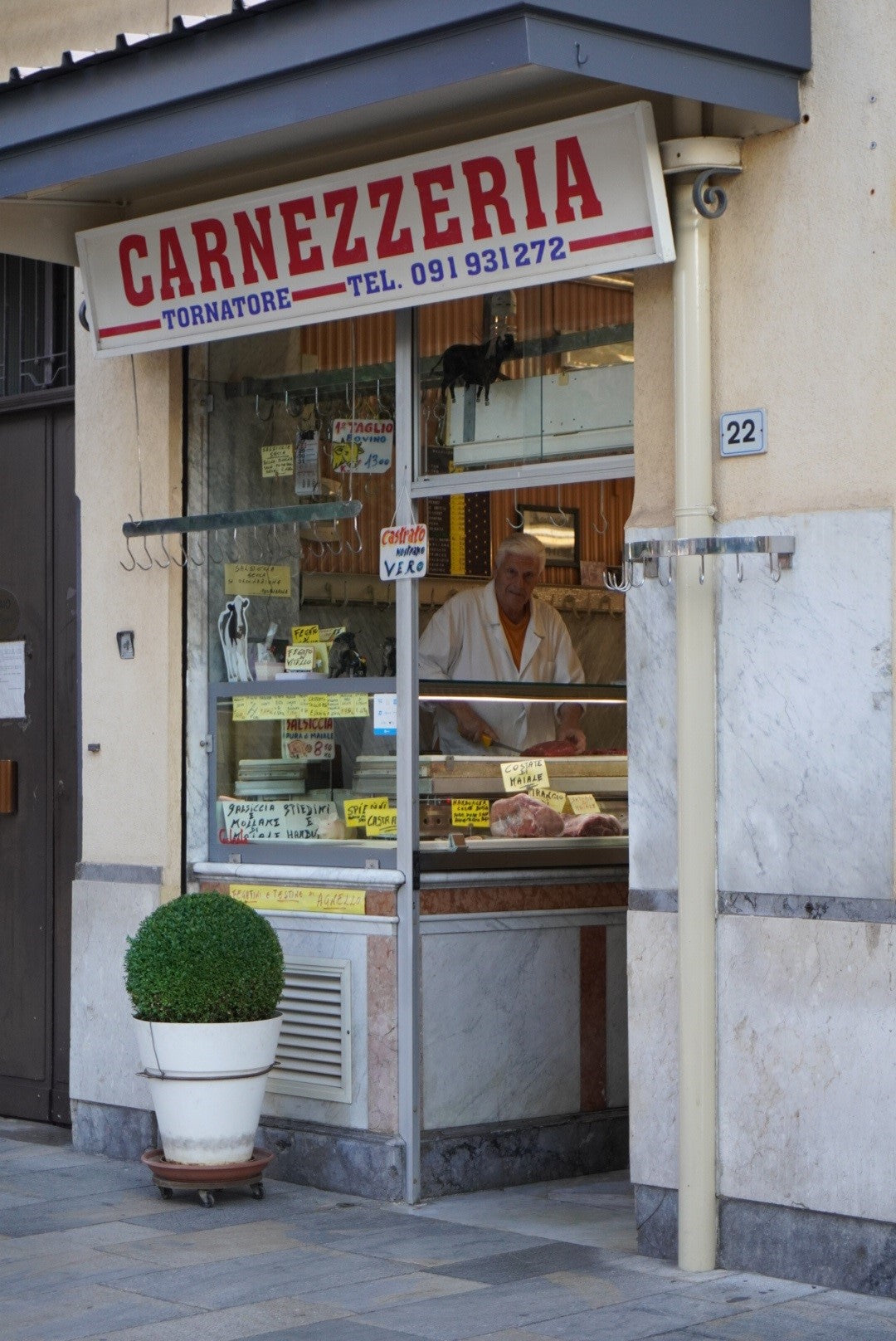 A butcher stands behind the counter inside a small Italian butcher shop called Carnezzeria Tornatore. The shop has a glass door, handwritten signs, and a potted plant outside by the entrance.