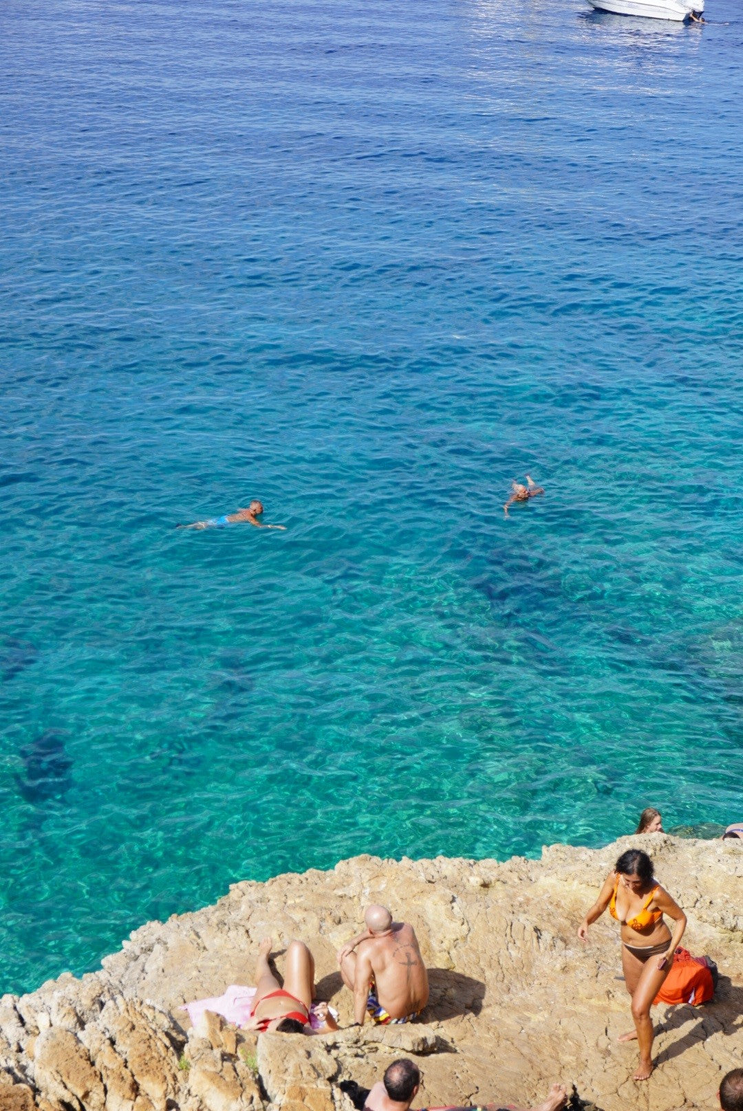 People relax on rocky shore while others swim in clear blue sea. One person stands by the water, another suns on a rock, and a boat floats in the distance under a sunny sky.