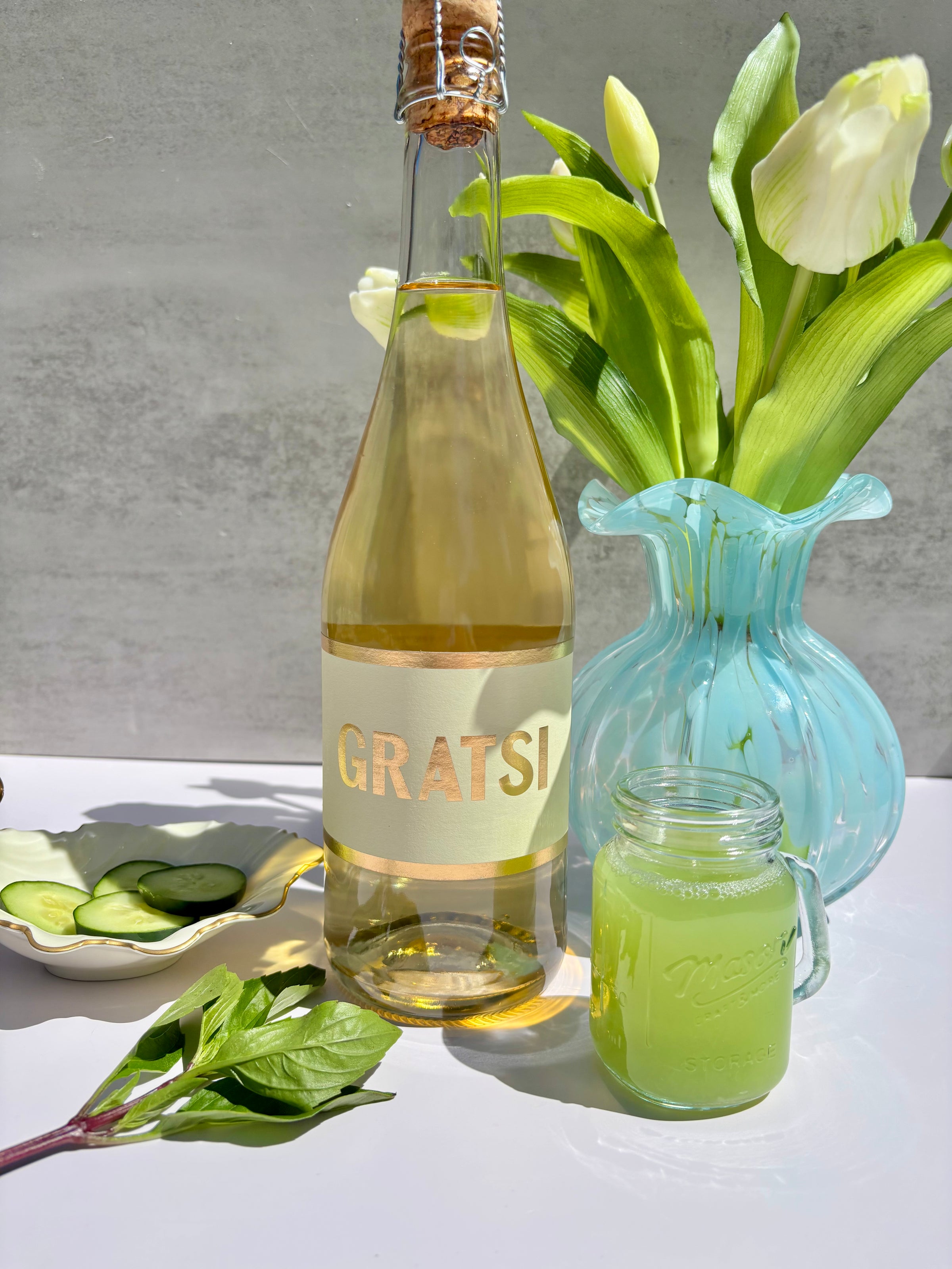 A bottle labeled GRATSI stands next to a glass jar of green juice, a blue vase with white tulips, a plate with cucumber slices, and fresh mint leaves, all arranged on a bright surface.