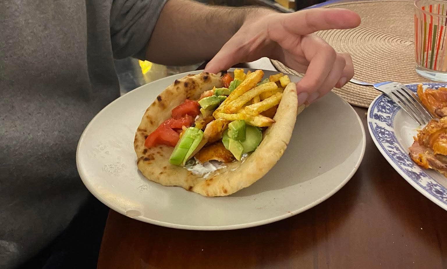 A person holds a pita filled with french fries, avocado, tomato, sauce, and other ingredients on a white plate, with another dish and a glass on the table.