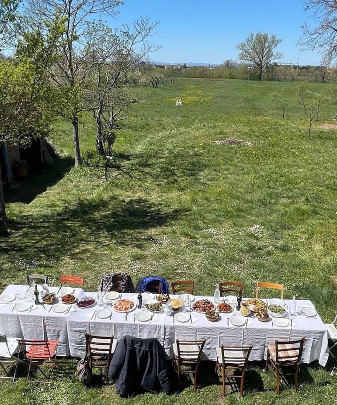 A long outdoor dining table is set with various dishes and plates, surrounded by mismatched chairs, on a grassy field under clear blue skies with trees and open landscape in the background.