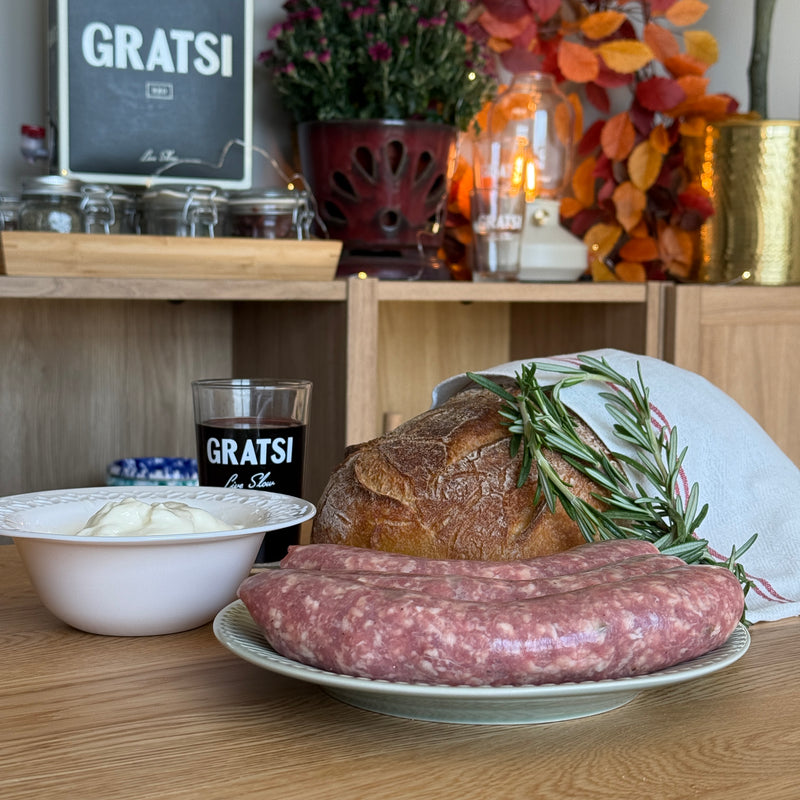 A rustic kitchen scene with a plate of raw sausage, a loaf of bread wrapped in a cloth with rosemary, a bowl of white sauce, and a glass labeled “GRATSI.” Autumn leaves and flowers decorate the background.