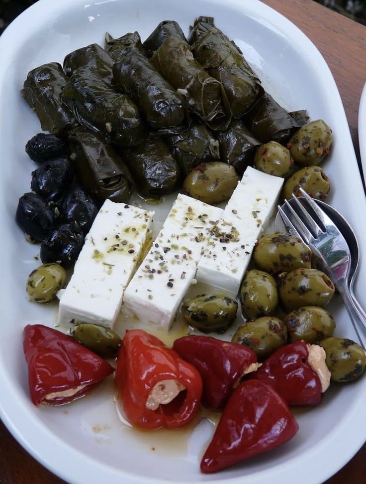 A white plate with stuffed grape leaves, black and green olives, cubes of feta cheese sprinkled with herbs, and red stuffed peppers. A small fork rests beside the olives.