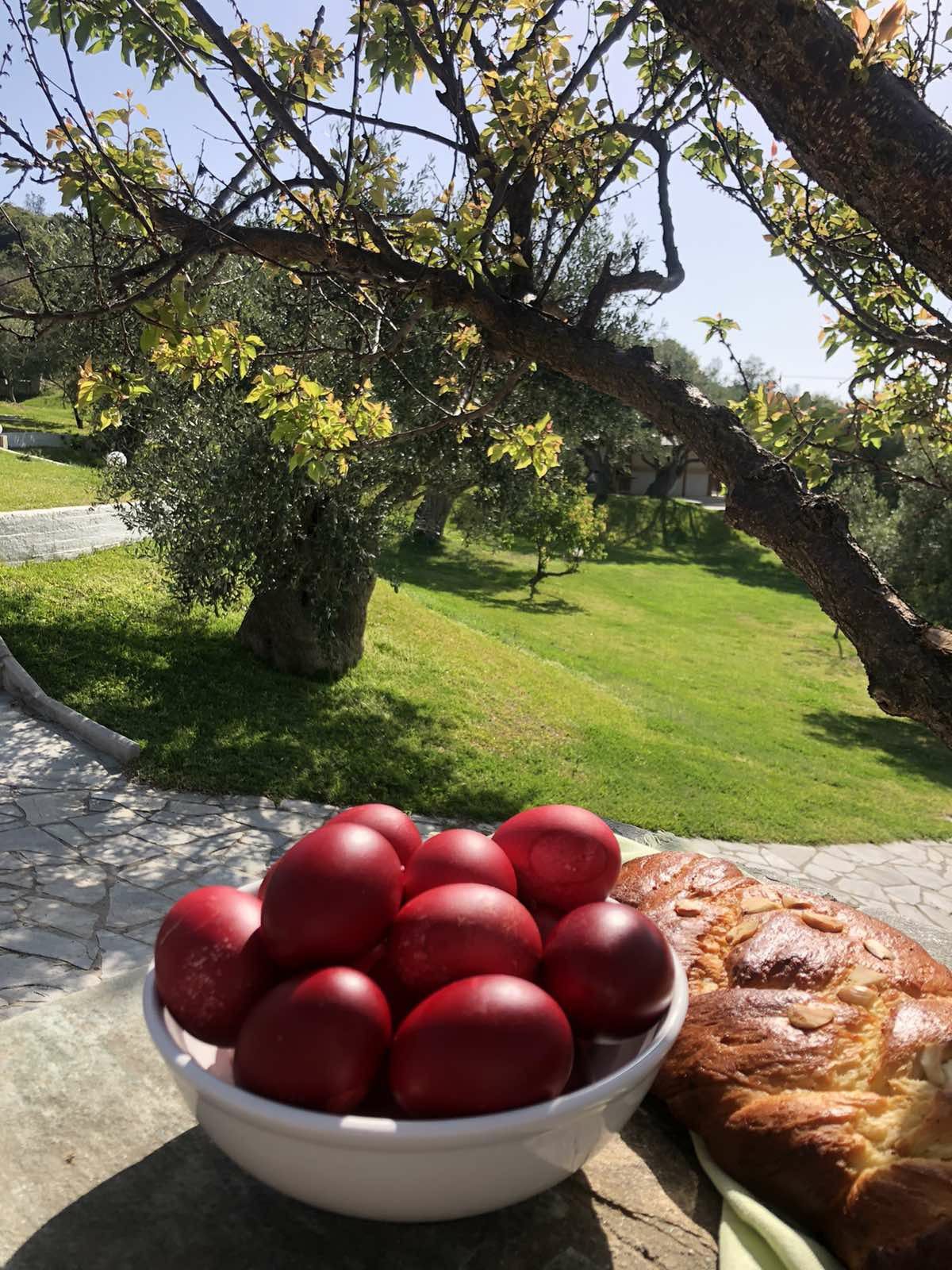 A bowl of red eggs and a loaf of bread sit on a stone table outdoors, with green grass, winding stone paths, and sunlit trees in the background.