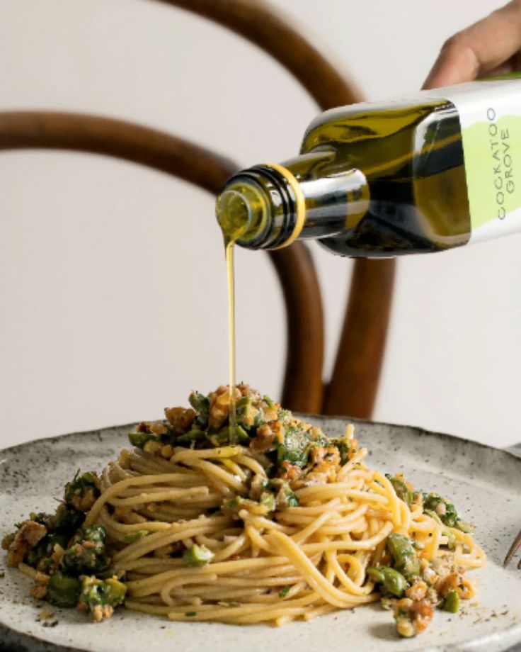 A hand pours olive oil from a bottle over a plate of spaghetti topped with chopped vegetables and herbs.