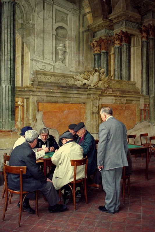 Seven elderly men, some seated and some standing, gather around a table playing cards in an ornate, historic room with tall columns, intricate architecture, and peeling walls. Wooden chairs surround the table.