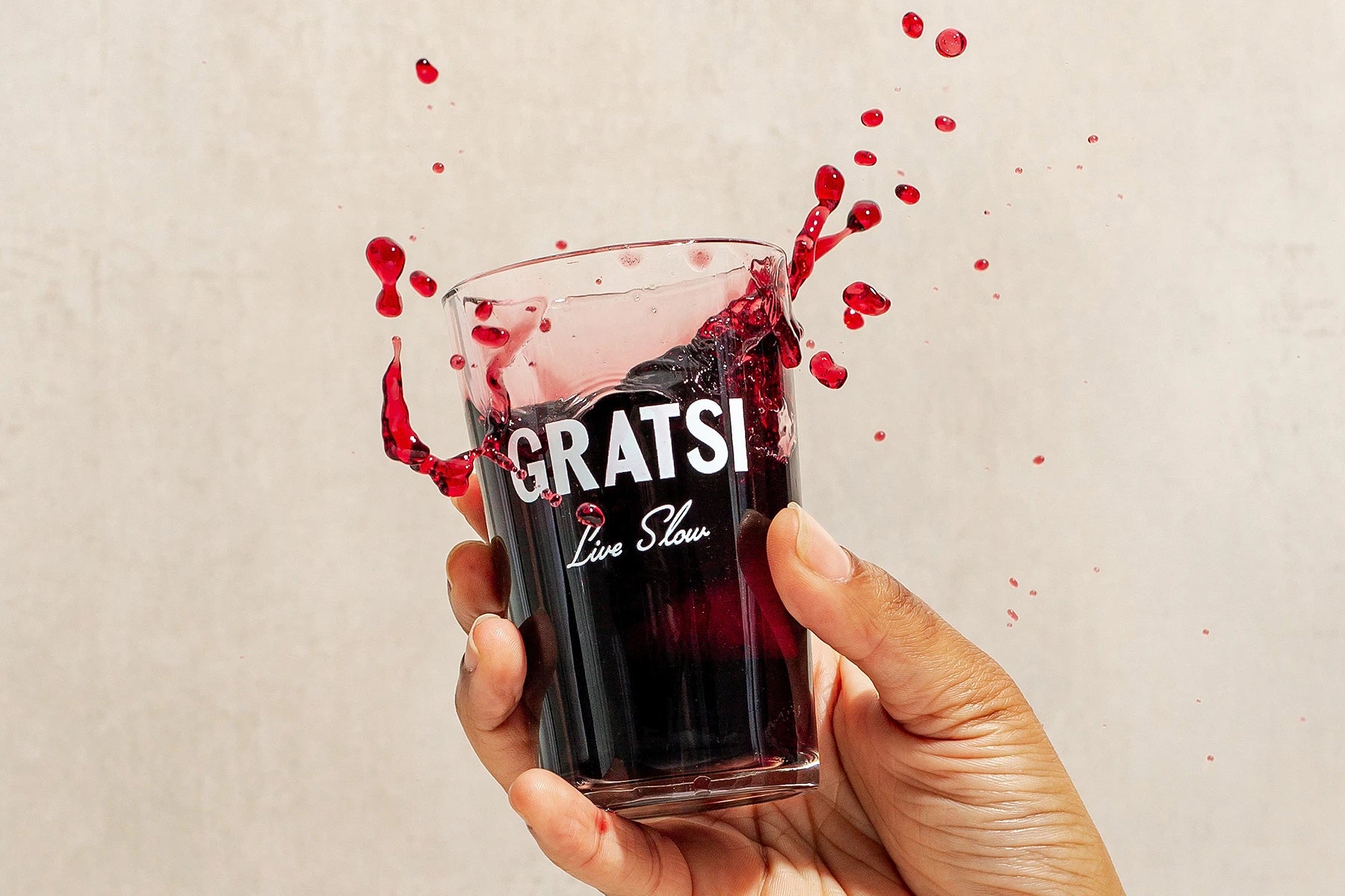 A hand holding a glass of red wine with the words GRATSI Live Slow as wine splashes out, frozen mid-air against a plain light background.