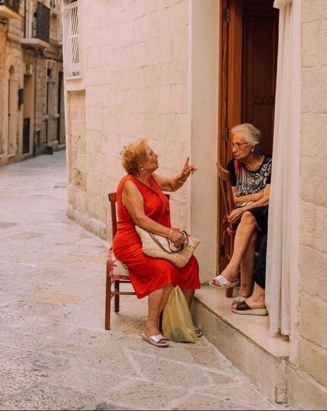 Two elderly women converse outside a stone building; one sits on a chair in a red dress gesturing, while the other sits in a doorway. The street is narrow and appears quiet and sunlit.