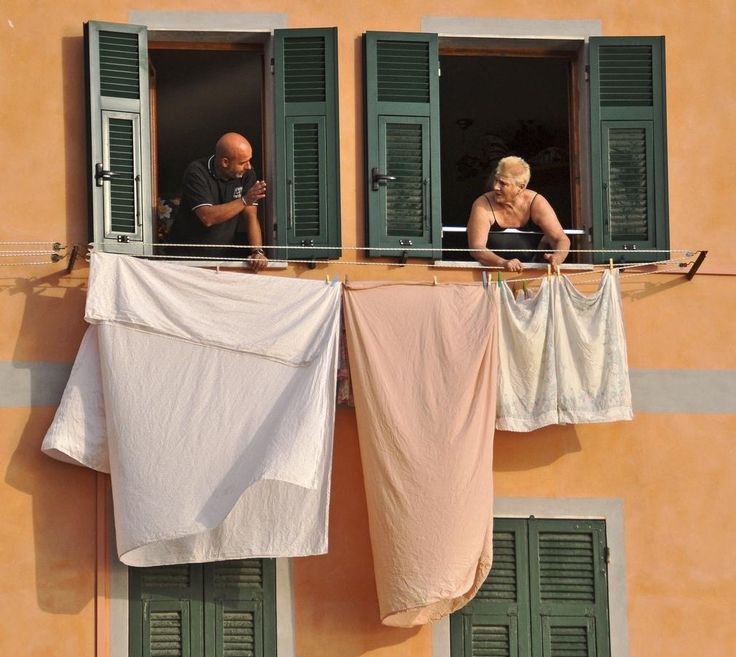 Two people leaning out of adjacent upper windows of a peach-colored building, chatting over laundry hanging on a line between them. The windows have green shutters, and large sheets are draped below.