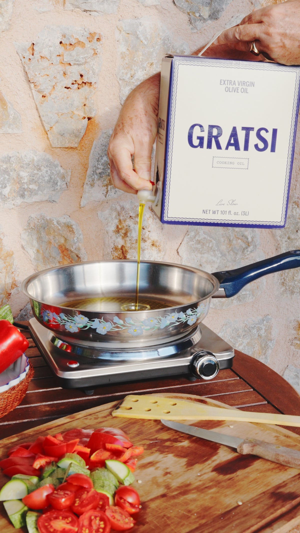 A person pours Gratsi extra virgin olive oil from a box into a floral-patterned frying pan on a portable electric burner.
