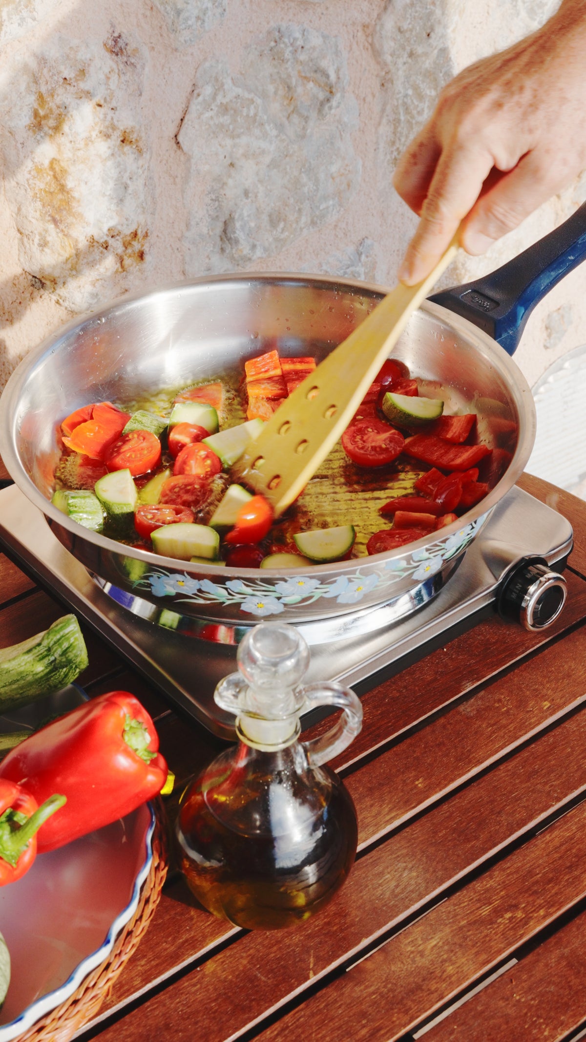 A hand uses a wooden spatula to stir sliced zucchini and red peppers in a stainless steel pan on a portable stove, with a bottle of olive oil and fresh vegetables nearby.