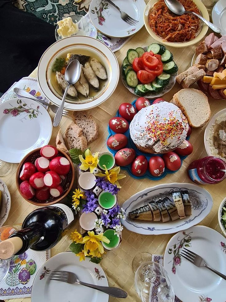 A festive table set with various dishes, including soup, sliced bread, fresh vegetables, red-dyed eggs, a decorated cake, fish, cold cuts, and a bottle of wine, with a bouquet of spring flowers in the center.