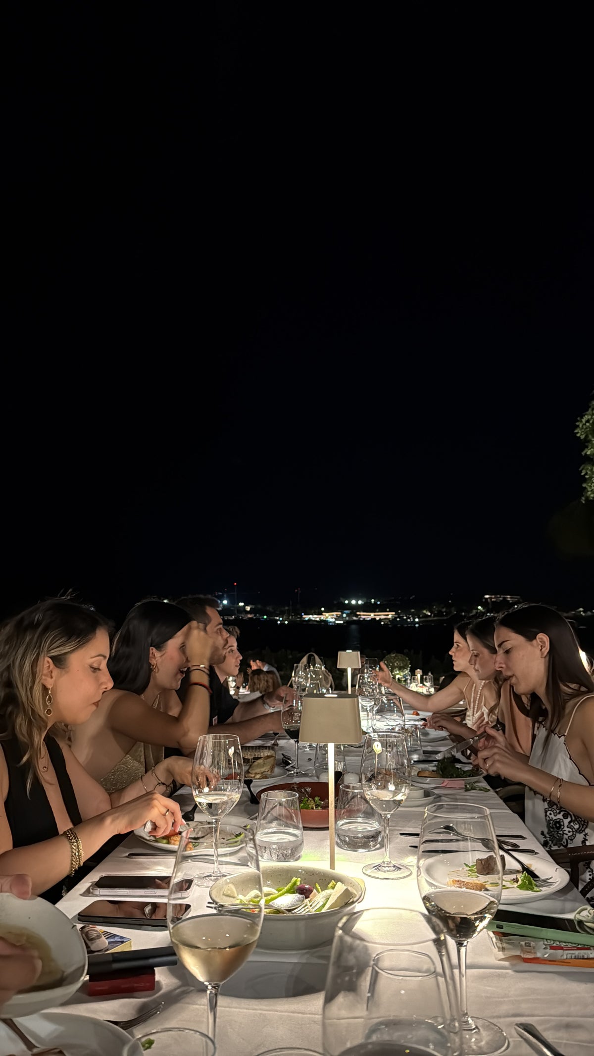 A group of people dine at a long, outdoor table at night, with city lights visible in the background. The table is set with plates, glasses, and candles.