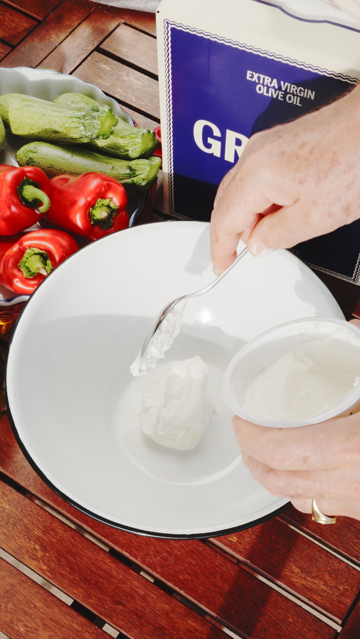 Hands using a spoon to add white yogurt or cheese from a container into a white bowl, with zucchini and red peppers in the background next to a tin of olive oil.