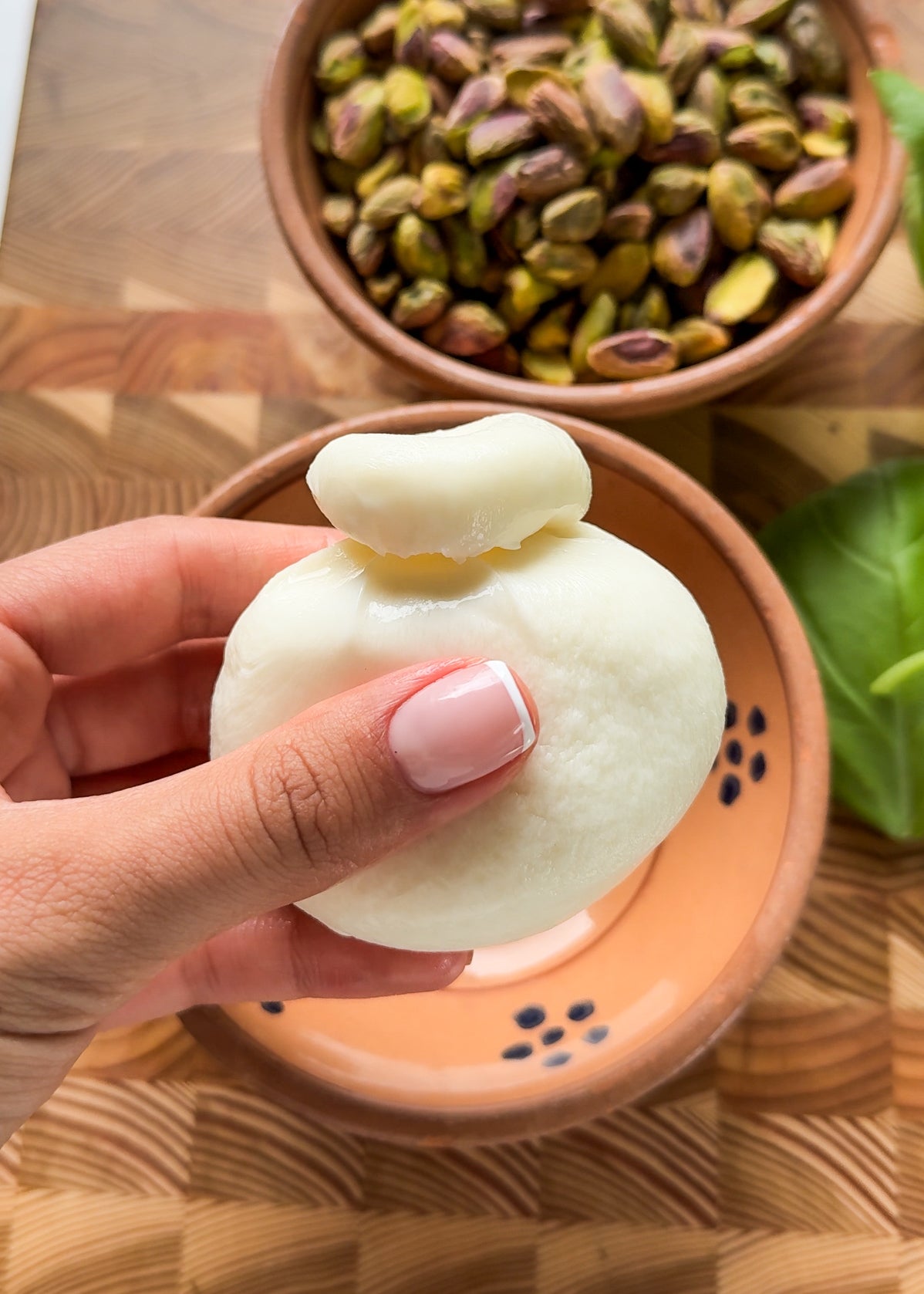 A hand with manicured nails holds a fresh ball of burrata cheese. In the background, theres a bowl of unshelled pistachios and a bowl with a leaf on a wooden surface.