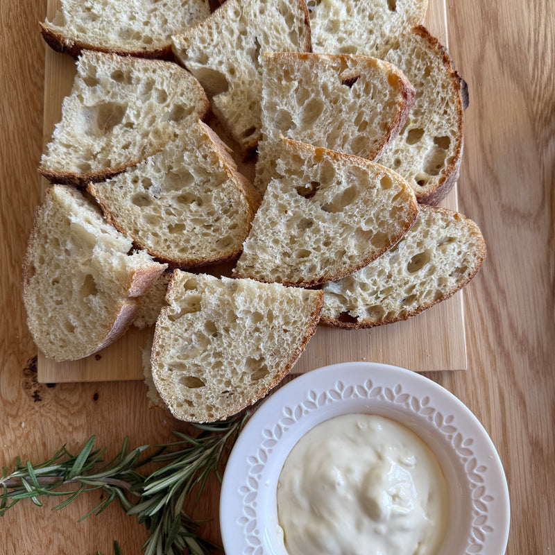 Slices of rustic bread are arranged on a wooden board next to a white bowl filled with creamy dip. A sprig of fresh rosemary lies nearby on the wooden surface.