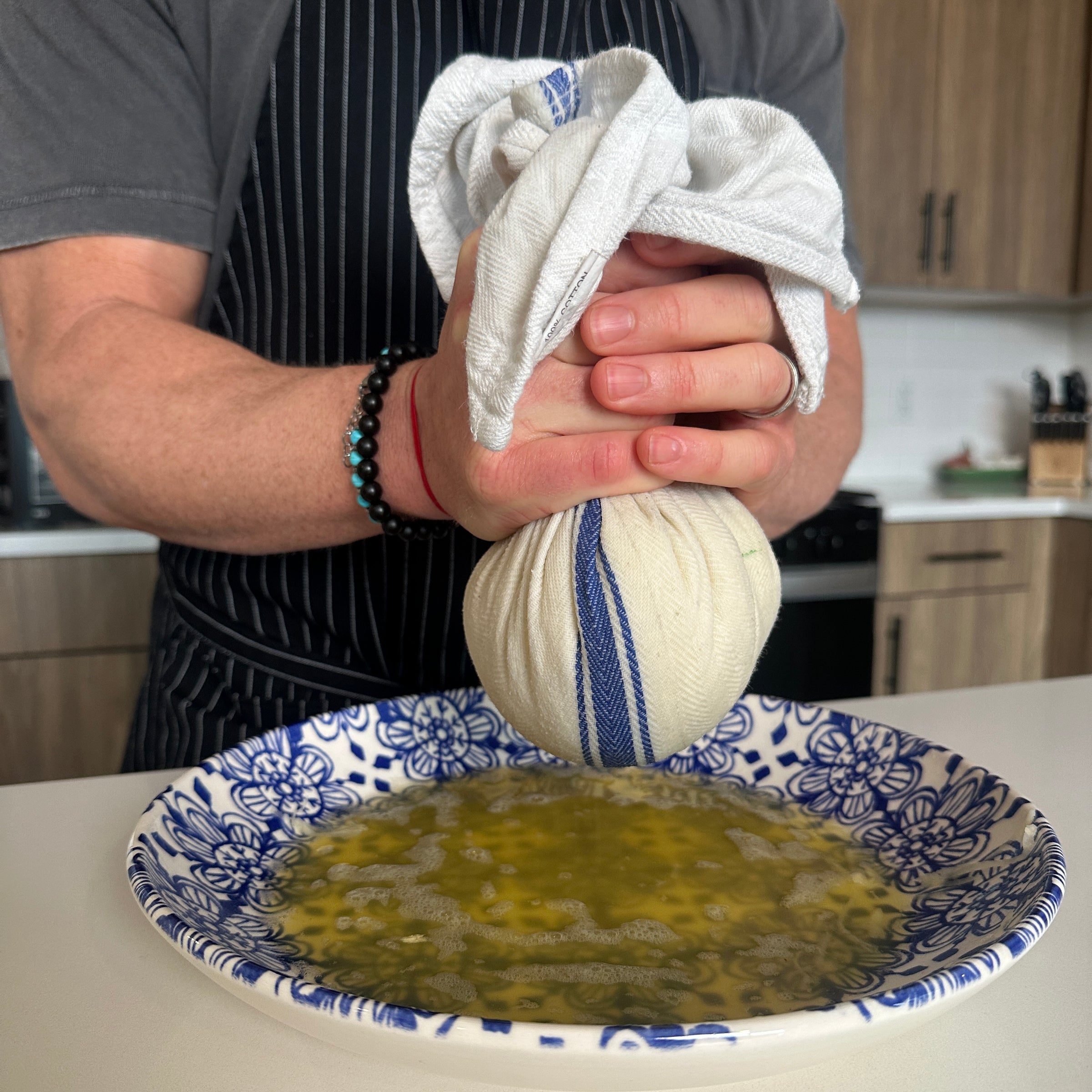 A person squeezes a cloth-wrapped bundle over a blue and white patterned bowl, extracting liquid. The scene takes place in a modern kitchen.