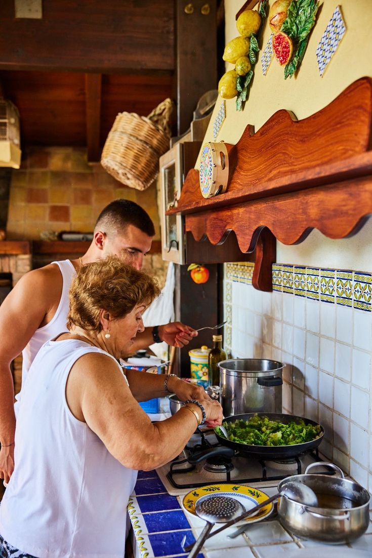 An older woman and a young man cook together in a cozy kitchen, stirring vegetables in a pan on the stove. Decorative tiles and hanging baskets adorn the walls.