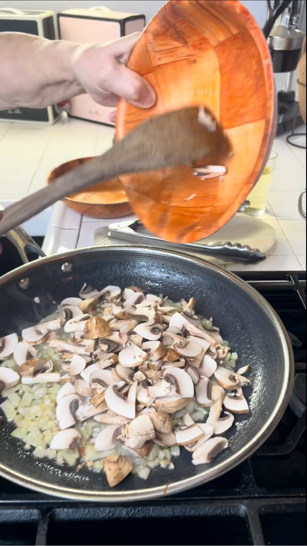 A hand pours sliced mushrooms from an orange bowl into a frying pan with chopped onions cooking on a stovetop. A wooden spoon stirs the mixture. Kitchen appliances and utensils are visible in the background.