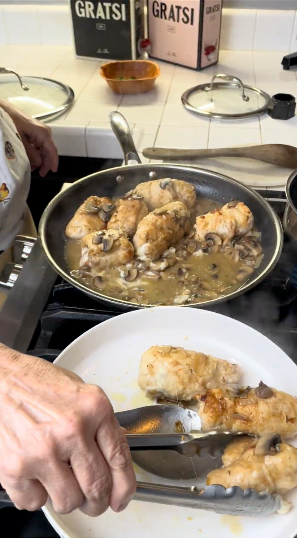 A person uses tongs to place breaded chicken pieces from a skillet with mushroom sauce onto a white plate on a stove. Two pots with lids and GRATSI boxes are in the background.