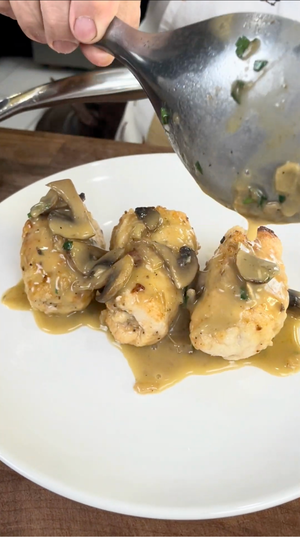 A hand pours creamy mushroom sauce from a large spoon over three pieces of stuffed chicken breast on a white plate.